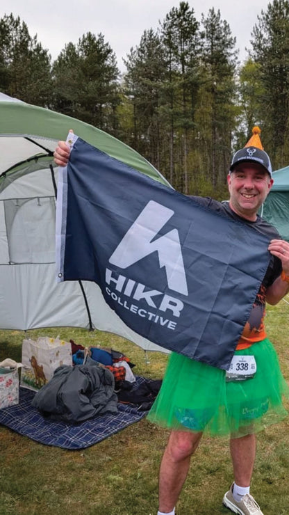 Person holding a HIKR Collective flag in front of a tent with trees in the background