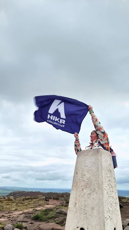 Person holding a flag with 'HIKR Collective' on top of a stone structure against a cloudy sky.