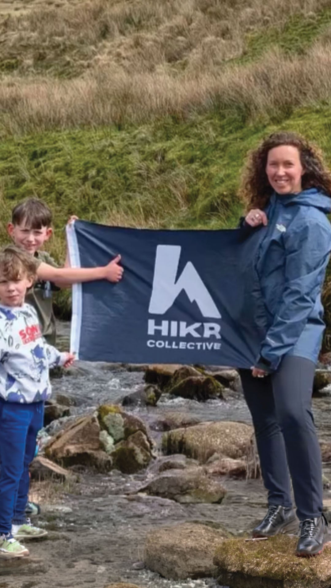 Two children and a woman holding a HIKR Collective flag in a natural setting.