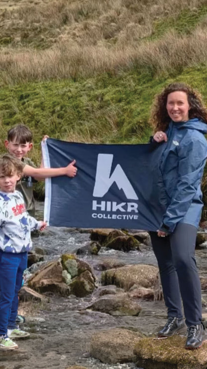 Two children and a woman holding a HIKR Collective flag in a natural setting.