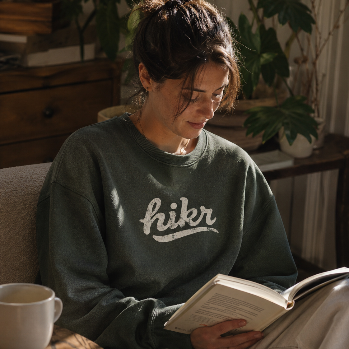 Person wearing a sweatshirt with 'hikr' text, reading a book in a cozy indoor setting.